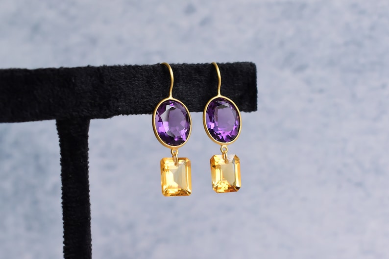 May include: A pair of gold earrings with a purple oval-shaped gemstone and a smaller, square-shaped yellow gemstone dangling below.