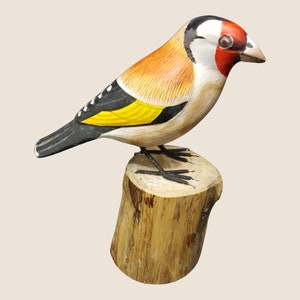 May include: Hand-painted wooden bird figurine of a goldfinch perched on a wooden stump. The bird is painted with realistic details, including black and white markings, a yellow breast, and a red head.