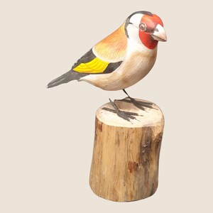 May include: A hand-carved wooden bird figurine of a goldfinch perched on a wooden stump. The bird is painted with realistic details, including a red head, black wings, and yellow breast.