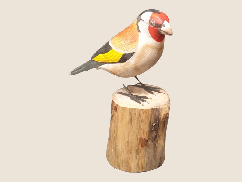 May include: A hand-carved wooden bird figurine of a goldfinch perched on a wooden stump. The bird is painted with realistic details, including a red head, black wings, and yellow breast.
