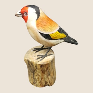 May include: Hand-carved wooden bird figurine of a goldfinch perched on a tree stump. The bird is painted with realistic details, including a red head, black wings, and yellow breast.