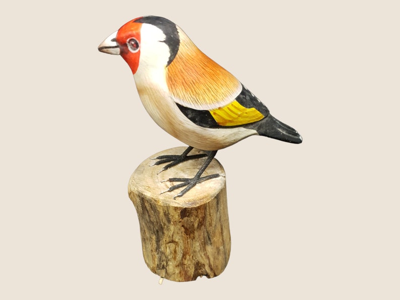 May include: Hand-carved wooden bird figurine of a goldfinch perched on a tree stump. The bird is painted with realistic details, including a red head, black wings, and yellow breast.