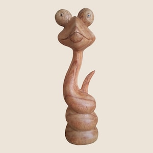 May include: A wooden sculpture of a snake with a long neck and large eyes. The snake is carved in a light brown colour and has a whimsical expression.