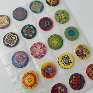 May include: A collection of colorful, circular pins with various geometric and abstract designs. The pins are in a clear plastic sheet, showcasing a variety of colors including blue, green, yellow, red, and purple. Each pin has a unique pattern.