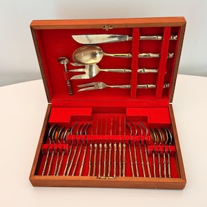 May include: A wooden storage box with a bright red velvet interior containing a set of gold-toned metal flatware. The utensils feature bamboo-style handles and include a knife, spoons, forks, and a small ladle arranged in molded slots.