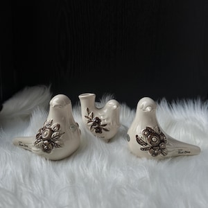 Puede incluir: Tres figuras de pájaros de cerámica crema con diseños florales marrones. Los pájaros están colocados sobre una superficie blanca y esponjosa. Cada pájaro tiene una forma y un patrón floral únicos, con el texto "Rosa Ljung" en la base.