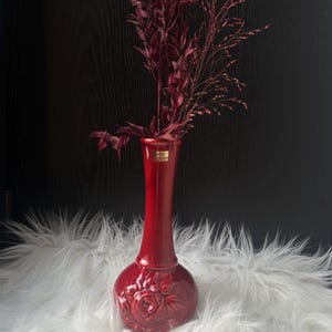 以下が含まれることがあります： 丸みを帯びたベースと細い首を持つ、赤いセラミックの花瓶。乾燥したバーガンディ色の花と草が活けられている。花瓶の底には花の模様があり、白いふわふわした表面に置かれている。