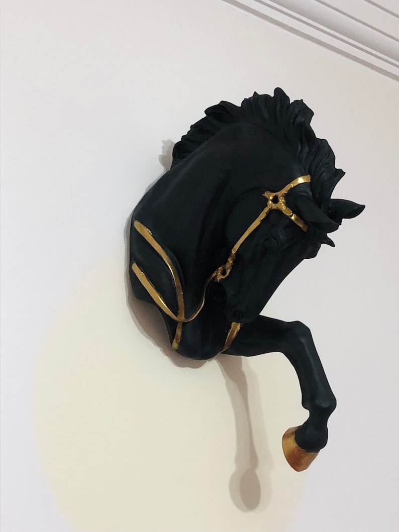 May include: Black horse head wall decor with gold accents. The horse's head is tilted to the side and the horse's mouth is slightly open. The horse's mane is flowing and the horse's tail is curled up.