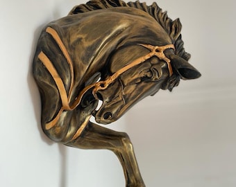 May include: A bronze-colored horse head sculpture with gold accents. The horse's head is tilted down and its leg is raised. The sculpture is mounted on a white wall.