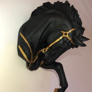 May include: Black horse head sculpture with gold accents. The horse's head is tilted to the side, and its mouth is slightly open. The sculpture is mounted on a wall.