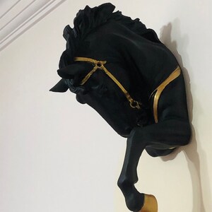 May include: Black horse head wall decor with gold accents. The horse's head is tilted to the side, and its mane is flowing. The horse's hoof is raised, and it is wearing a gold bridle.
