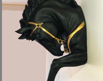 May include: Black horse head wall sculpture with gold accents. The horse's head is tilted to the side, and its mane is flowing. The horse is wearing a gold bridle.