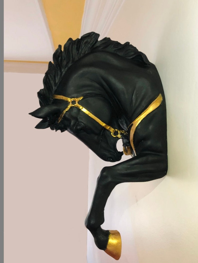 May include: Black horse head wall sculpture with gold accents. The horse's head is tilted to the side, and its mane is flowing. The horse is wearing a gold bridle.