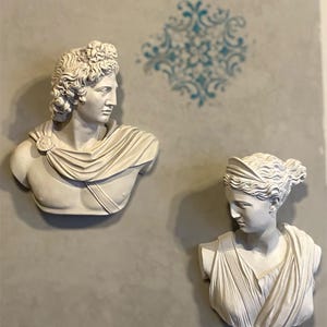 Könnte beinhalten: Zwei weiße Gipsbüsten klassischer Figuren sind an einer grauen Wand angebracht. Die Büste links zeigt einen Mann mit lockigem Haar und einem drapierten Gewand. Die Büste rechts zeigt eine Frau mit hochgestecktem Haar und einem drapierten Gewand. Ein blaues Blumendesign befindet sich im Hintergrund.