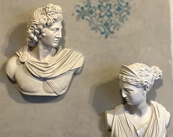 May include: Two white plaster busts of classical figures are mounted on a gray wall. The bust on the left depicts a man with curly hair and a draped garment. The bust on the right shows a woman with her hair up and wearing a draped garment. A blue floral design is in the background.