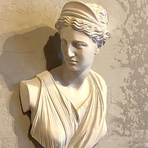 以下が含まれることがあります： 古典的な特徴を持つ女性の白い石膏胸像。彫刻は、波状の髪、ヘアバンド、ドレープされた服を着た女性を描いています。胸像はテクスチャード加工された壁に取り付けられており、滑らかな彫刻と粗い背景のコントラストを生み出しています。