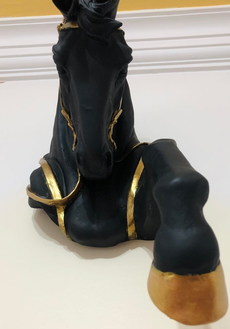 May include: Black horse head sculpture with gold accents. The horse is lying down with its head raised. The sculpture is made of a smooth, black material and has a glossy finish.