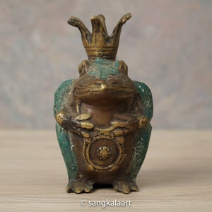 以下が含まれることがあります： 王冠をかぶったブロンズ製のカエルの置物。緑と金の風合いで、カエルは座って胸に装飾的なエンブレムを持っています。装飾品または収集品である可能性があります。