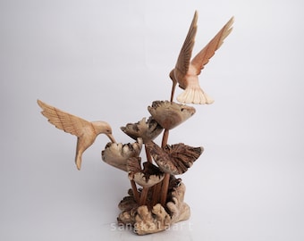 May include: A wooden sculpture of two hummingbirds hovering near a cluster of stylized flowers. The birds are carved with detailed wings and beaks, and the flowers have a textured, natural appearance. The sculpture is made of light brown wood.