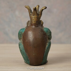 以下が含まれることがあります： 王冠をかぶったブロンズ製のカエルの置物。カエルは茶色の体で、側面と脚にターコイズのアクセントがあります。王冠は明るいブロンズ色です。置物は木製の表面に置かれています。