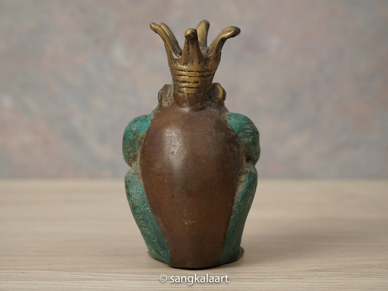 以下が含まれることがあります： 王冠をかぶったブロンズ製のカエルの置物。カエルは茶色の体で、側面と脚にターコイズのアクセントがあります。王冠は明るいブロンズ色です。置物は木製の表面に置かれています。