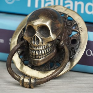 May include: A detailed brass skull door knocker with a ring handle. The skull is centered on a decorative circular backplate. The knocker has an aged, antique appearance. The background includes blue books.