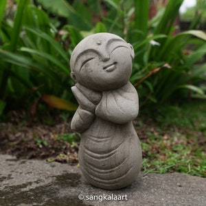 May include: A small, gray stone statue of a smiling Buddha with hands clasped in prayer. The statue is standing on a gray stone surface.