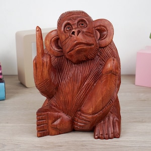 May include: A hand-carved wooden sculpture of a monkey, with reddish-brown tones. The monkey is seated and giving the middle finger with its right hand. The sculpture has detailed carving and a natural wood grain.