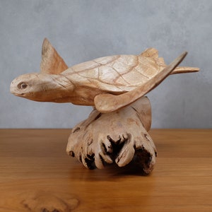 Puede incluir: Una escultura de tortuga marina de madera tallada a mano con un acabado de madera natural. La tortuga está posada sobre un trozo de madera a la deriva.
