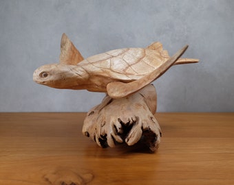 Puede incluir: Una escultura de tortuga marina de madera tallada a mano con un acabado de madera natural. La tortuga está posada sobre un trozo de madera a la deriva.