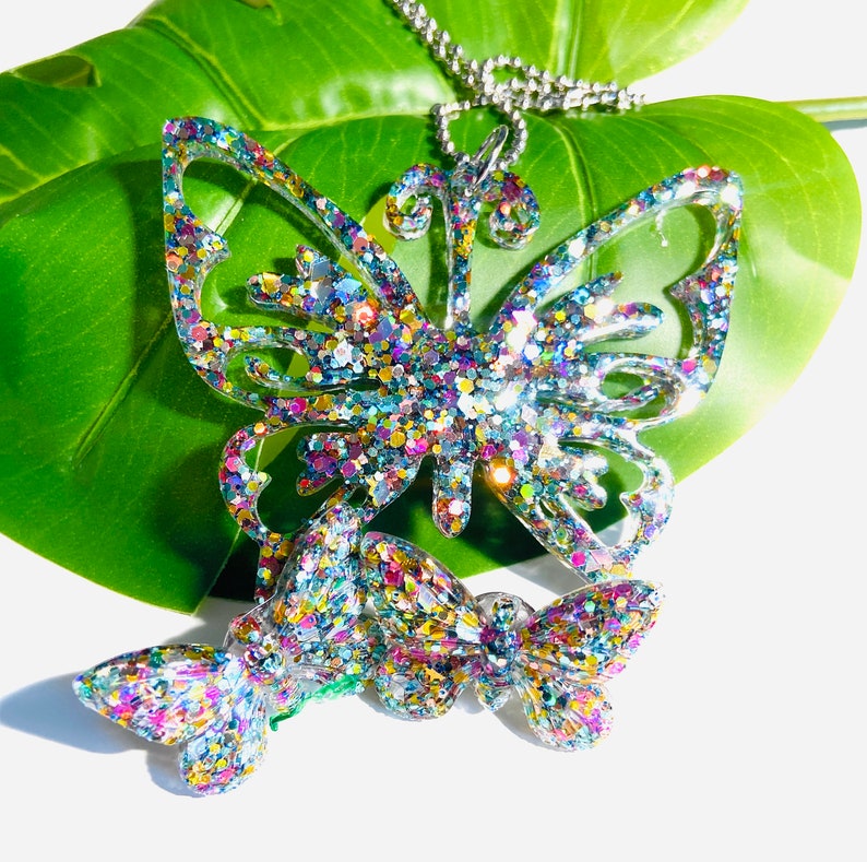 Puede incluir: Un colgante de collar que presenta una forma de mariposa grande e intrincada y un dije de mariposa m&aacute;s peque&ntilde;o, ambos hechos de resina transparente incrustada con part&iacute;culas de purpurina multicolor reflectante, suspendidos de una cadena de bolas plateada.