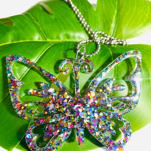 Puede incluir: Un colgante con forma de mariposa hecho de resina transparente rellena de purpurina reflectante multicolor. El colgante presenta detalles de alas recortadas y est&aacute; sujeto a un collar de cadena de bolas de metal plateado.
