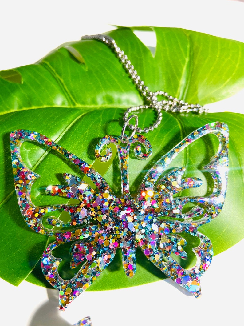 Puede incluir: Un colgante con forma de mariposa hecho de resina transparente rellena de purpurina reflectante multicolor. El colgante presenta detalles de alas recortadas y est&aacute; sujeto a un collar de cadena de bolas de metal plateado.