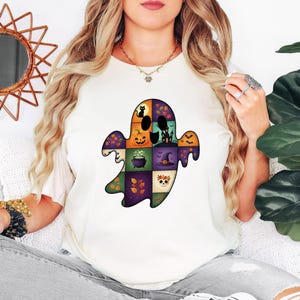 May include: Cream-colored t-shirt featuring a colorful ghost design. The ghost is composed of various Halloween-themed squares, including a black cat, jack-o'-lantern, and witch's hat. The design is a patchwork of orange, purple, green, and black.