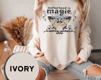 May include: A long-sleeved, ivory colored t-shirt with a butterfly graphic and the text "motherhood is magic".