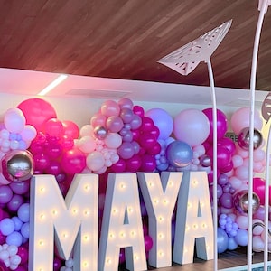 An indoor pool party event setup for a sweet sixteen birthday featuring large light-up marquee letters spelling "MAYA" with a backdrop of pink and purple balloons. Heart-shaped pink pool floats are placed in the pool for decoration.