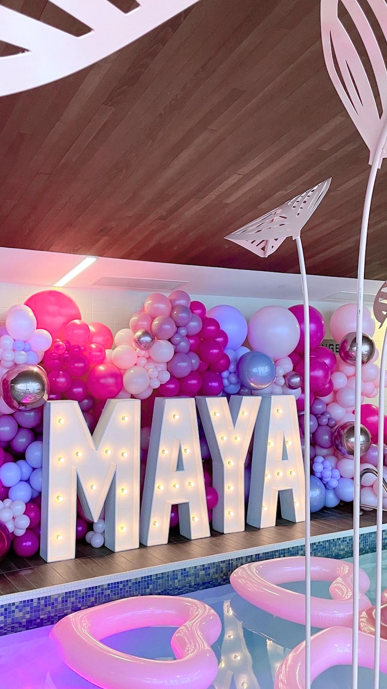 An indoor pool party event setup for a sweet sixteen birthday featuring large light-up marquee letters spelling "MAYA" with a backdrop of pink and purple balloons. Heart-shaped pink pool floats are placed in the pool for decoration.