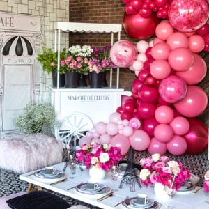 May include: A pink and white floral themed party setup with a white flower cart, a pink and white balloon arch, and a white wall with a black and white cafe sign that says "Drianna's Cafe".