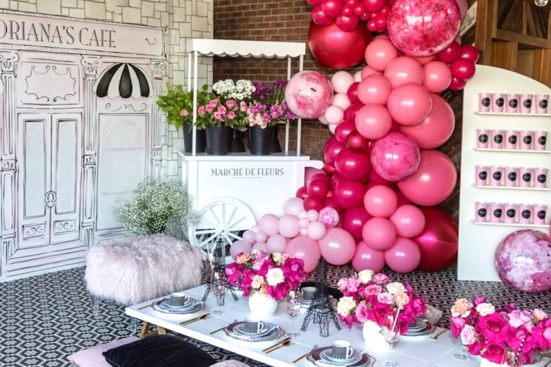 May include: A pink and white floral themed party setup with a white flower cart, a pink and white balloon arch, and a white wall with a black and white cafe sign that says "Drianna's Cafe".
