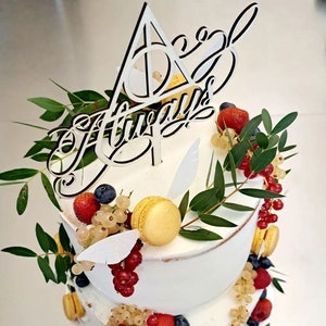 Könnte beinhalten: Eine dreistöckige weiße Hochzeitstorte, verziert mit frischem Obst, Macarons und Grünpflanzen. Der Tortendecker ist ein Holzschild mit der Aufschrift "Always" und einem Symbol der Heiligtümer des Todes.