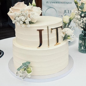 Könnte beinhalten: Ein zweistöckiger, weiß glasierter Kuchen, verziert mit frischen Rosen, Schleierkraut und Grünzeug. Goldfarbene Initialen "JIT" schmücken die obere Etage. Der Kuchen steht auf einer weißen Kuchenplatte, ideal für eine Hochzeit oder Feier.