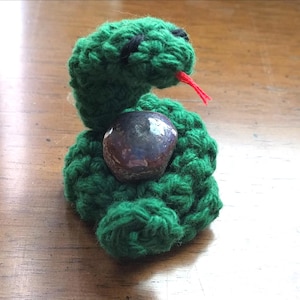 May include: A handmade green crochet snake with black eyes and a red tongue. The snake is holding a dark, speckled stone. The craft project is displayed on a wooden surface.