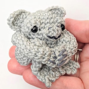Może przedstawiać: Ręcznie robiony szydełkowy miś koala w odcieniach szarości. Amigurumi koala ma czarne oczy i mały czarny nos. Widoczne są ramiona i nogi koali, a on sam wydaje się obejmować. Ściegi szydełkowe są wyraźnie zdefiniowane.