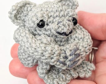 Op de afbeelding: Een handgemaakte gehaakte koala beer in grijstinten. De amigurumi koala heeft zwarte veiligheidsoogjes en een kleine zwarte neus. De armen en benen van de koala zijn zichtbaar en hij lijkt zichzelf te omarmen. De haaksteken zijn duidelijk gedefinieerd.