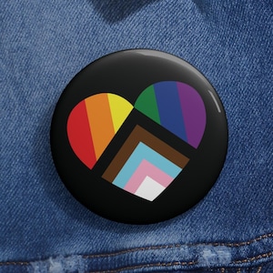 May include: A black button with a rainbow heart design. The heart is made up of different colored stripes, including red, orange, yellow, green, blue, indigo, and violet. The heart also includes a brown stripe with a blue, pink, and white stripe on top of it.