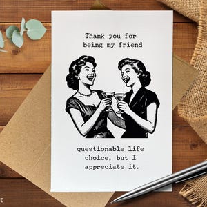 May include: A white greeting card featuring a black ink illustration of two people laughing and holding cocktail glasses. The text reads: Thank you for being my friend, questionable life choice, but I appreciate it. The card has a textured paper finish.