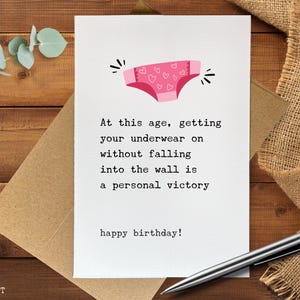 May include: A white greeting card featuring a pink pair of underwear with a heart pattern at the top. The text reads: At this age, getting your underwear on without falling into the wall is a personal victory. happy birthday!