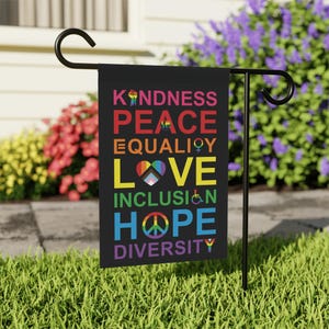May include: A black garden flag with the words "KINDNESS, PEACE, EQUALITY, LOVE, INCLUSION, HOPE, DIVERSITY" in rainbow colors. Each word has a symbol associated with it, such as a peace sign, a heart, and a raised fist.