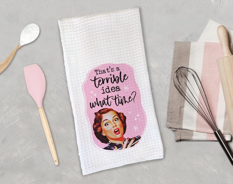 May include: White kitchen towel with a pink border and a retro-style illustration of a woman with the text "That's a terrible idea. What time?"