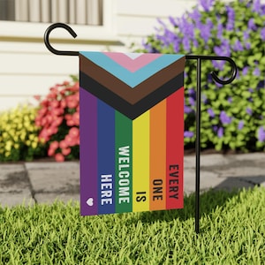 May include: A garden flag with a rainbow pride flag design and the text "Every one is welcome here" in white lettering. The flag is attached to a black metal pole.